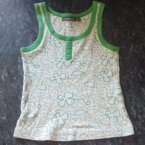 Grön och vit linnetopp Race Marine - Snygg linnetopp från Race Marine i vitt med grönt mönster och gröna kanter. Toppen har tre knappar framtill och ett lekfullt mönster av prickar och blommor. Perfekt för varma dagar och enkel att matcha med shorts eller jeans.