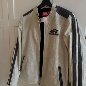 Skinnjacka från Diesel - Riktigt snygg vit skinnjacka från Diesel med svarta axelpartier och ränder längs ärmarna. Jackan har perforerat skinn, dragkedja framtill och coola Diesel-loggor både fram och bak. Perfekt för dig som vill sticka ut med en sportig och edgy look. Är endast använd 1 gång. NYPRIS: 8000kr