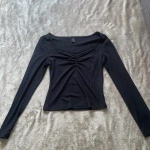 Svart ribbad topp från Romwe (SHEIN) - Svart långärmad topp från Romwe med ribbad struktur och snygg rynkdetalj framtill. Toppen har en v-ringad hals med diskret spetskant och sitter figurnära. Perfekt att styla med jeans eller kjol för en trendig look.