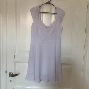 Ljuslila kortklänning med volang - Supersöt vit  kortklänning från Dame med v-ringning och breda axelband. Klänningen har volangkant nertill och är sydd i ett tunt, flowy material som ger en romantisk vibe. Perfekt för sommarens alla tillfällen! Köparen betalar frakten 