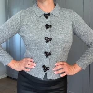 Grå kofta med pärlrosetter från Zara - Supersöt grå kofta från Zara med långa ärmar, två fickor framtill och dekorativa svarta pärlrosetter som knäppning. Koftan är ribbstickad och har en klassisk krage som ger en chic vibe. Perfekt för dig som gillar unika detaljer och vill sticka ut lite extra.