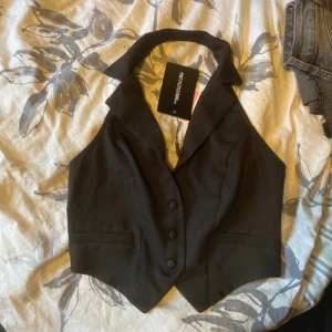 Svart väst från PrettyLittleThing - Snygg svart väst med krage och knappar framtill från PrettyLittleThing. Västen har en figurnära passform, två fuskfickor och djup v-ringning. Perfekt att styla över en skjorta eller t-shirt för en trendig look.