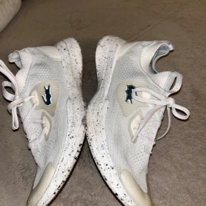 Vita sneakers från Lacoste med stickad ovandel - Snygga vita sneakers från Lacoste med stickad mesh-ovandel och klassisk krokodil-logga på sidan. Skorna har snörning, platt sula med svart stänkmönster och förstärkt tåparti i beige. Perfekta för en clean och sportig look.