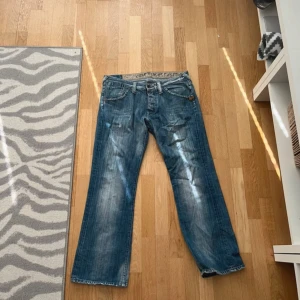 Blå bootcut jeans med slitningar - Säljer ett par blå jeans med bootcut passform och snygga slitningar. Jeansen har klassisk femficksdesign, normal midja och är tillverkade i denim. Perfekta för dig som gillar en avslappnad och retro stil.