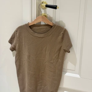 Beige stickad kortärmad tröja - En enkel och stilren beige stickad tröja med korta ärmar och rund hals. Tröjan har ribbade muddar vid ärmslut och nederkant, vilket ger en snygg och avslappnad look. Perfekt att matcha med jeans eller kjol för en clean vibe. står ingen storlek men skulle säga att det är som en xxs!