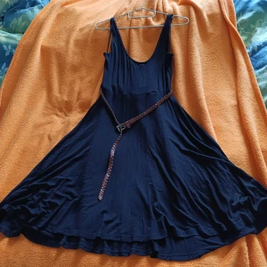 Svart knälång klänning med flätat bälte - Enkel och snygg svart klänning med djup urringning både fram och bak. Klänningen har ett brunt flätat bälte i midjan som ger en cool kontrast. Tillverkad i ett mjukt och lätt tyg, perfekt för varma dagar eller när du vill känna dig extra fri.