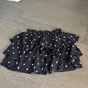 Svart volangkjol med stjärnor H&M - Supersöt svart kortkjol från H&M med vita stjärnor och flera volanger. Kjolen har bred, smockad midja som är stretchig och sitter skönt. Tillverkad i mjuk viskosblandning som känns lätt och luftig. Perfekt för dig som gillar en lekfull och trendig stil.