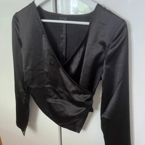 Svart omlottopp i satin från Gina Tricot - Snygg svart omlottopp från Gina Tricot i glansigt satinmaterial. Toppen har lång ärm, v-ringning och en cool asymmetrisk nederkant som ger en trendig look. Perfekt för dig som gillar stilrena och moderna plagg med det lilla extra.