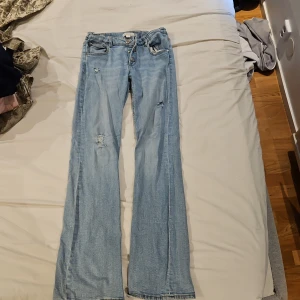 Ljusblå bootcut jeans Gina Tricot - Snygga ljusblå lågmidjade  jeans från Gina Tricot i bootcut-modell. Jeansen har slitna detaljer och hål på benen för en trendig look. Klassisk femficksdesign, knappgylf och tillverkade i mjukt denimtyg. Perfekta för dig som gillar en avslappnad och cool stil.