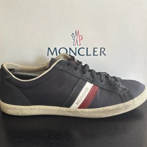 Moncler sneakers i marinblå mocka - Snygga sneakers från Moncler i marinblå mocka och textil med vita och röda detaljer på sidan. Skorna har snörning, rund tå och en vit, något vintage-inspirerad sula. Moncler-logga på sidan och plös. Väldigt bra skick 