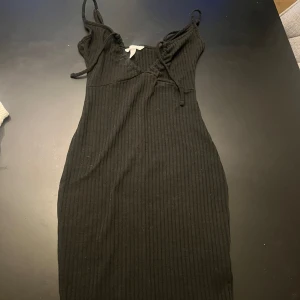 Svart ribbad kortklänning med snörning - Svart ribbad kortklänning med smala axelband och snygg snörning framtill vid bysten. Klänningen är figurnära och har en enkel, stilren look som passar perfekt till sneakers eller boots. Materialet känns mjukt och stretchigt för en bekväm passform.