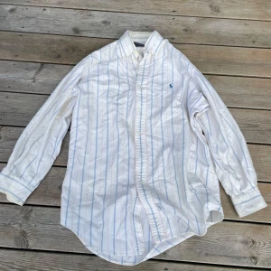 Vit randig skjorta från Polo Ralph Lauren - Vit skjorta från Polo Ralph Lauren med tunna blå och röda ränder. Klassisk button-down krage, långärmad och med broderad logga på bröstet. Tillverkad i bomull och har en avslappnad passform. Perfekt för en clean och stilren look.