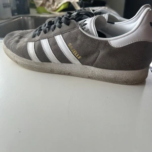 Adidas Gazelle grå/vita sneakers - Säljer ett par klassiska Adidas Gazelle sneakers i grå mocka med vita detaljer och mörkgråa snören. Perfekta för dig som gillar retrostil och snygg streetwear.