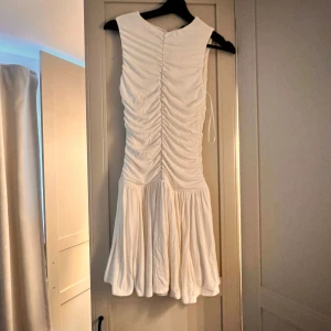 Vit kortklänning med rynkade detaljer - Supersöt vit kortklänning med rynkade detaljer längs hela livet och en volangig kjol. Ärmlös modell med rund halsringning, perfekt för sommarens alla tillfällen. Klänningen har en feminin och fräsch vibe som är lätt att styla.