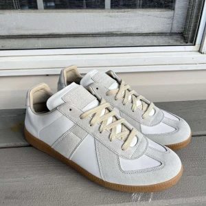 Vita Maison Margiela sneakers - Stilrena sneakers från Maison Margiela i vitt skinn och mocka med beige snörning och klassisk gummisula. Snygg minimalistisk design med diskreta detaljer och rund tå. Perfekta för dig som gillar clean och modern stil.