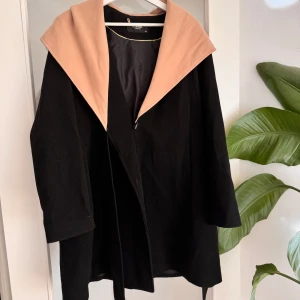 Svart kappa med beige/ljusrosa krage - Stilren svart kappa från LC Waikiki med bred, beige krage som ger en snygg kontrast. Jackan har lång ärm och snörning runt midian. Perfekt för dig som gillar klassisk men trendig look. 