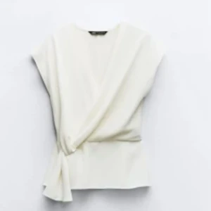 Vit omlottopp från Zara - Säljer en stilren vit omlottopp från Zara i storlek M. Toppen har v-ringning, knyt i sidan och är ärmlös med en lätt glansig finish. Perfekt för dig som gillar en clean och chic look med snygg passform. Annvänd 4-5 gånger så i nyskick