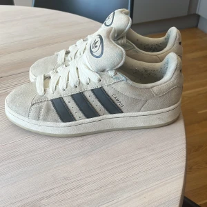 Beige adidas Campus sneakers mocka - Snygga adidas Campus sneakers i beige mocka med klassiska svarta ränder på sidorna och vit sula. Skorna har rund tå, platt sula och breda vita snören. Ikonisk adidas-logga på plösen. Perfekta för dig som gillar retrostil och streetwear.