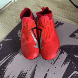 Nike Phantom Vision röda fotbollsskor - Snygga Nike Phantom Vision fotbollsskor i rött med svarta och silvriga detaljer. Skorna har hög krage i stretchigt material och mönstrad ovandel för extra grepp. Sulan har fasta dobbar för spel på gräs. Perfekta för dig som vill sticka ut på planen.