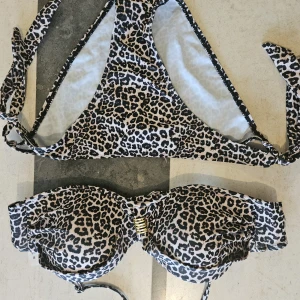 Leopardmönstrad bikini med snörning - Snygg bikini i leopardmönster med svarta, bruna och beige toner. Överdelen har vadderade kupor, gulddetalj framtill och knyt i ryggen. Underdelen har klassisk trosa med knyt i sidorna. Materialet är stretchigt i polyester och elastan – perfekt för stranden!