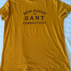 Gul Gant t-shirt med tryck - Gul t-shirt från Gant med svart tryck på bröstet där det står 'New Haven Connecticut'. Klassisk rund hals och korta ärmar. Perfekt för dig som gillar en sportig och avslappnad stil.