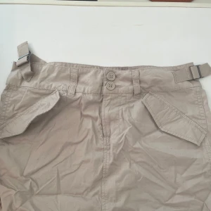 Beige utility kortkjol med fickor - Snygg beige utility-inspirerad kortkjol med stora fickor fram och bak, dubbla knappar i midjan och justerbara spännen i sidorna. Perfekt för en avslappnad och cool look. Gjord i bomull för en skön känsla.