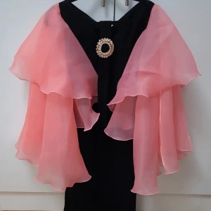 Svart klänning med rosa volangcape - Unik svart långklänning med dramatiska, skira rosa volanger över axlarna och en stor glittrig brosch framtill. Klänningen har en elegant och iögonfallande look, perfekt för dig som vill sticka ut. Materialet känns lätt och flowy.