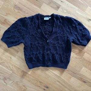 Mörkblå stickad kofta från Kappahl - Supersöt mörkblå stickad kofta från Kappahl med korta ärmar och v-ringning. Koftan har ett unikt hålmönster och knäpps med tre knappar framtill. Perfekt att slänga över en topp för en chill look.