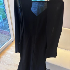 Svart långklänning - Elegant svart långklänning med v-ringad hals. Den har en mantel över armarna - du trär igenom armen och den lägger sig fint på utsidan av armen. Klänningen har en figurnära passform upptill och är sydd i ett slätt tyg som faller snyggt. Den har en bred ”svans”.