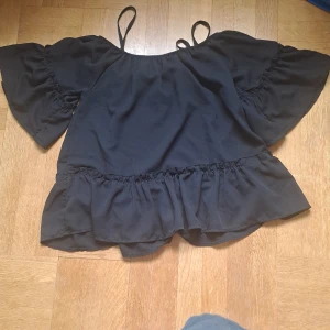 Svart volangtopp  - Svart topp från Myholi med volangkant nertill och vida, korta ärmar. Toppen har smala axelband och en luftig passform. 🩷.