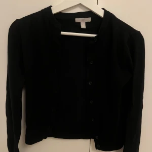 Svart kofta från H&M med knappar - En klassisk svart kofta från H&M med rund halsringning och knäppning framtill. Långärmad och i mjukt material som passar perfekt till lager-på-lager. Enkel och stilren design som funkar till det mesta.