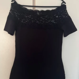 Svart spets topp Jacqueline de Yong - Supersnygg svart topp från Jacqueline de Yong med offshoulder och bred spets upptill. Toppen har korta ärmar och en figurnära passform som ger en stilren look. Perfekt för dig som gillar en enkel men elegant stil.