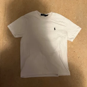 Vit t-shirt från Polo Ralph Lauren - Klassisk vit t-shirt från Polo Ralph Lauren med korta ärmar och rund hals. Diskret broderad logga i svart på bröstet. Tillverkad i mjuk bomull för en skön och avslappnad stil. Perfekt till jeans eller shorts för en clean look.