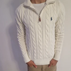 Vit kabelstickad tröja Polo Ralph Lauren - Vit kabelstickad tröja från Polo Ralph Lauren med halv dragkedja och hög krage. Klassisk broderad logga på bröstet i mörkblått. Tröjan är i mjuk ullblandning och har långa ärmar med ribbade muddar. Perfekt för en clean och stilren look.