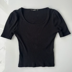 Svart ribbad topp från Bik Bok - Svart ribbad topp från Bik Bok med rundad, djup urringning och korta puffärmar. Sitter snyggt och har en tight passform som framhäver figuren. Perfekt att styla med jeans eller kjol för en clean look