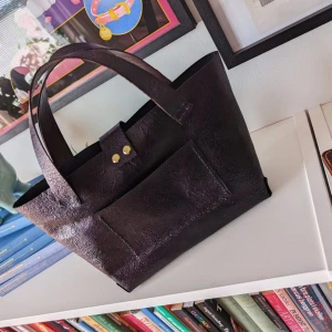 skinnväska med ytterfack - Stilren väska i skinn med rejäla handtag och ett praktiskt ytterfack. Väskan har en enkel, boxig form och stängs med en rem och guldfärgad knapp. Perfekt för dig som gillar minimalistisk design och vill ha plats för allt du behöver. Skimrande mörklila färg med vackert mönster. Handsydd av mig