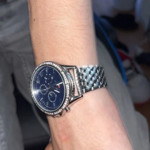Tommy Hilfiger klocka med blå urtavla - Snygg klocka från Tommy Hilfiger med blå urtavla, tre små urtavlor och silverfärgat armband i metall. Runt boetten sitter glittrande stenar som ger extra bling. Klassisk design med Hilfiger-logga på spännet och urtavlan.