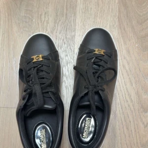Svarta sneakers från Michael Kors - Stilrena svarta sneakers från Michael Kors i skinn med vita sulor och svarta snören. Snygg guldfärgad MK-detalj framtill och logga även på sulan. Perfekta för dig som gillar en clean look med exklusiv känsla. De är använda ett par fåtal gånger då de är för små men annars är de i jättebra skick☺️
