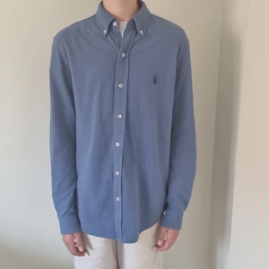 Blå skjorta från Polo Ralph Lauren - Snygg blå skjorta från Polo Ralph Lauren med klassisk button-down krage och broderad logga på bröstet. Skjortan har vita knappar och är långärmad, tillverkad i mjuk bomull. Perfekt för dig som gillar stilren och enkel look. Modellen på bilden är 170 och väger 53. 