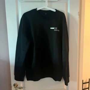 Svart sweatshirt med tryck THE ENDING - Svart sweatshirt med rund hals och vitt tryck på bröstet där det står 'THE ENDING'. Tröjan har långa ärmar och en klassisk, avslappnad passform. Perfekt för en chill och stilren look.