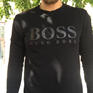 Svart långärmad tröja Hugo Boss - Svart långärmad tröja från Hugo Boss med stor BOSS-logga framtill. Enkel och stilren design med rund halsringning. Perfekt för dig som gillar klassiska streetwear-vibbar och vill ha en tröja som sticker ut med sitt tydliga tryck.