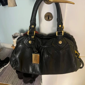 Svart handväska Marc by Marc Jacobs - Säljer en svart handväska från Marc by Marc Jacobs med snygga guldfärgade metalldetaljer och en stor metallbricka framtill. Väskan är gjord i mjukt skinn och har dubbla handtag med coola detaljer. Perfekt för dig som vill ha en klassisk men ändå edgy väska.