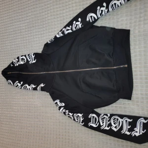 Svart bomberjacka med tryck - Svart bomberjacka med stora vita tryck i gotisk stil längs ärmar och huva. Jackan har dragkedja framtill, två fickor och ribbade muddar. Snygg streetwear-vibe och perfekt för dig som gillar statement-plagg.