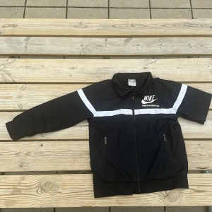 Svart Nike vindjacka med vita detaljer - Svart vindjacka från Nike med vita ränder över bröst och ärmar. Jackan har dragkedja framtill, två fickor med dragkedja och meshfoder. Klassisk Nike-logga och texten 'There is no finish line' på bröstet. Perfekt för en sportig look.