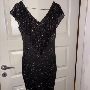 Svart glittrig långklänning med volang - Supersnygg svart långklänning med glittrigt tyg och v-ringning. Klänningen har volangdetalj över axlarna och en figurnära passform. Perfekt för dig som vill sticka ut med lite extra glitter.
