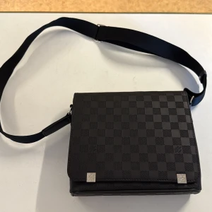 Louis Vuitton axelväska i svart skinn - Säljer en snygg svart axelväska från Louis Vuitton med klassiskt rutigt Damier-mönster. Väskan har justerbar axelrem, silverfärgade metalldetaljer och är tillverkad i exklusivt skinn. Perfekt för dig som vill ha en stilren och ikonisk accessoar.