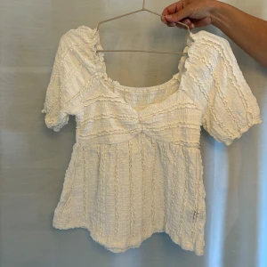 Vit blus med volangdetaljer - Helt ny Supersöt vit blus med puffiga korta ärmar och volangdetaljer över hela toppen. Den har en lätt rynkad struktur och en söt, romantisk känsla med rundad halsringning. Perfekt för dig som gillar en feminin och trendig stil.