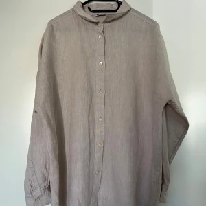 Beige linneskjorta, Cubus, L - Beige linneskjorta från Cubus med långa ärmar och knappar framtill. Skjortan har en krage och manschetter med knappar. Lös passform. Nyskick. 100% linne.   Mått:  Längd ca 70 cm Byst ca 60 cm Midja ca 60 cm Ärmlängd ca 50 cm Axel till axel ca 65 cm