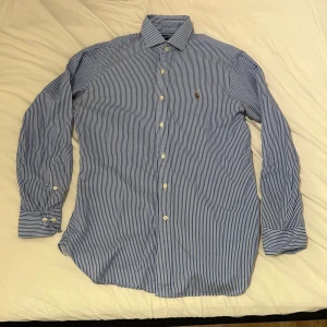 Blåvit randig skjorta Polo Ralph Lauren - Snygg blå och vit randig skjorta från Polo Ralph Lauren i slim fit-modell. Skjortan har klassisk krage, långa ärmar med manschett och knäppning framtill. Broderad logga på bröstet. Perfekt för dig som gillar stilrena och tidlösa plagg.