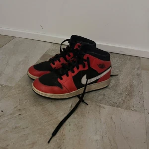 Nike Air Jordan 1 Mid röd/svart 38 - Säljer ett par Nike Air Jordan 1 Mid i klassisk röd och svart färg med vita swoosh-detaljer. Skorna har snörning, rund tå och platt sula. Tillverkade i skinn och syntet, perfekt för dig som gillar streetstyle och ikoniska sneakers.